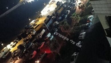 Nighttime city street with many parked cars along both sides and bright lights reflecting off water to the left.",