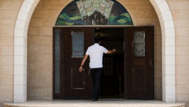 Man in a white shirt walks through open double doors of a stone building, under a colorful stained-glass arch window.