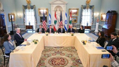 Formal meeting around a U-shaped table in a decorated room with flags (US and Israel) behind the panelists, including diplomats and translators photographed by journalists.