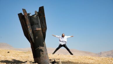 Person in a white shirt and dark pants jumps with arms outstretched beside a large, rusted metal structure in a desert under a clear blue sky.