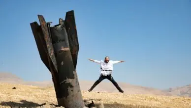 Person in a white shirt and dark pants jumps with arms outstretched beside a large, rusted metal structure in a desert under a clear blue sky.