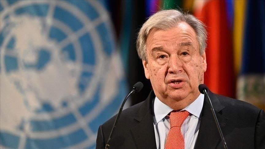 Elderly man in a dark suit and coral tie speaks at a podium with two microphones, in front of a UN backdrop and multicolored flags.