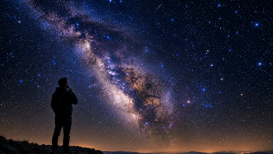 Silhouette of a person standing on a rocky hill at night, gazing at a bright Milky Way across a starry sky.